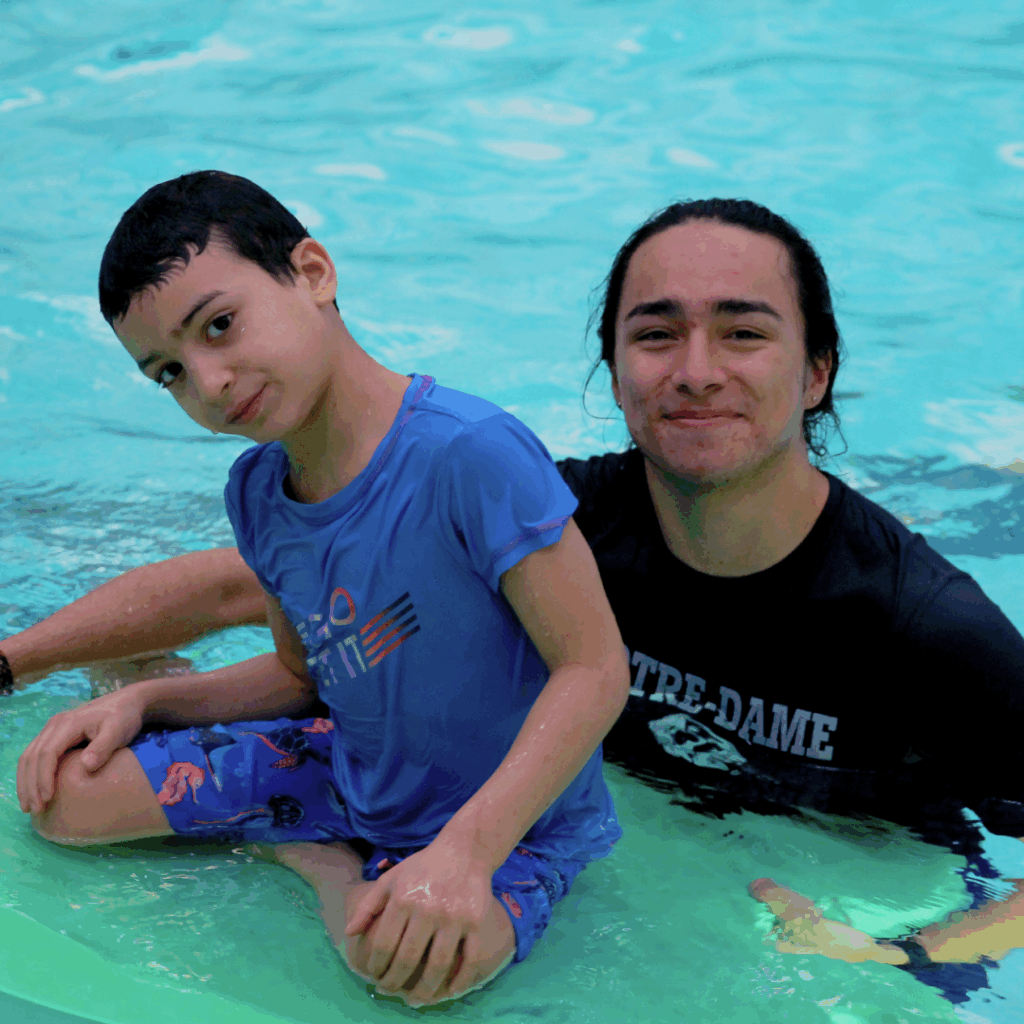 Jeune heureux assis sur une planche flottante dans la piscine avec son accompagnateur souriant qui le soutien.