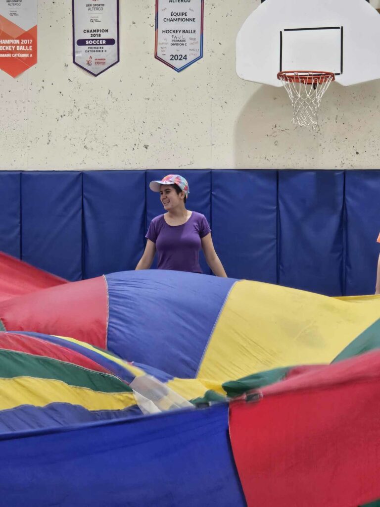 Jeune debout dans un gymnase, secouant un énorme parachute coloré (jaune, vert, bleu et jaune). 