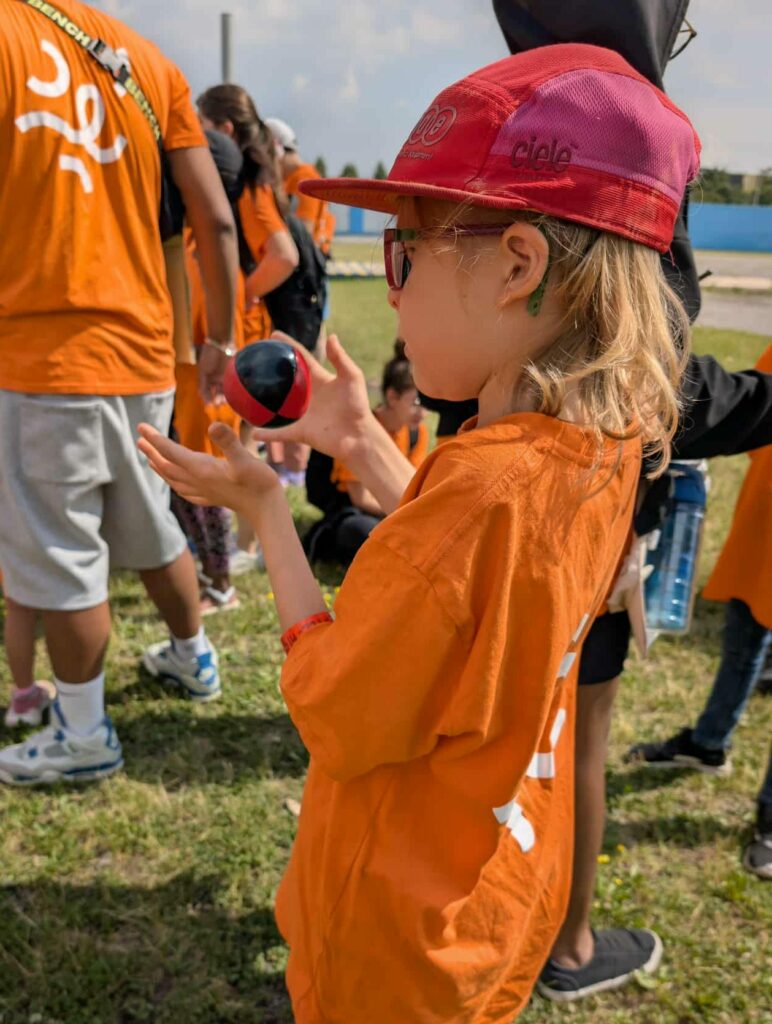 Jeune debout à l'extérieur dans la pelouse, lançant une balle de jonglerie dans les air, avec d'autres jeunes à l'arrière.