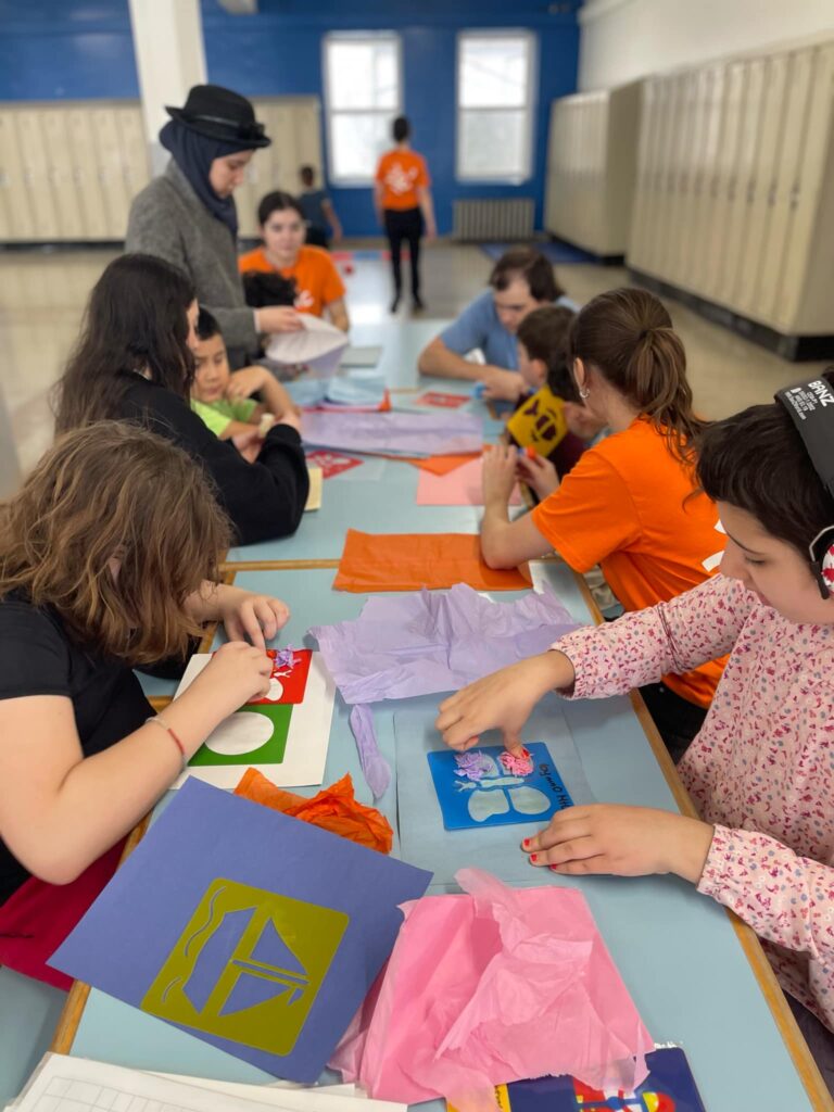 Plusieurs jeunes assis face à face sur une table de cafétéria, faisant des activités de coloriage et bricolage.