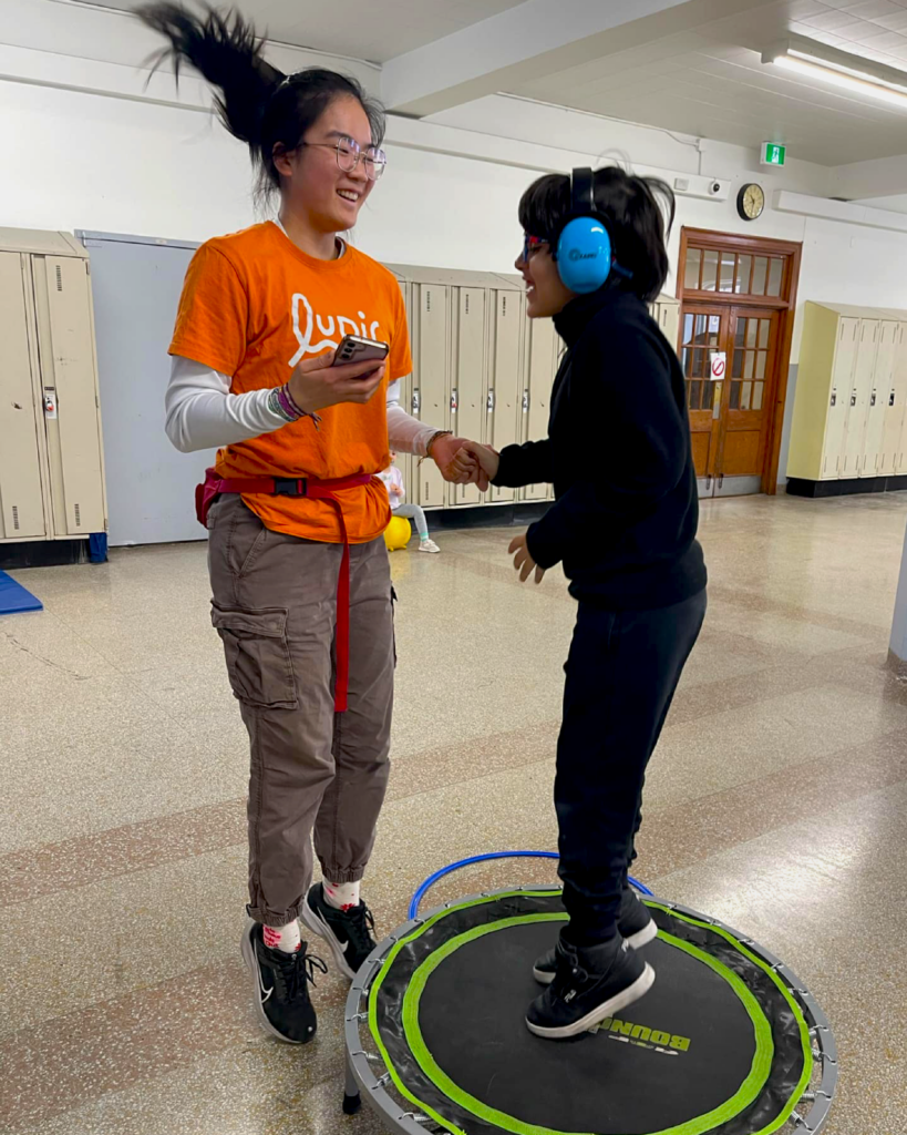 Accompagnatrice souriante tenant la main d'un jeune qui saute sur un petit trampoline.