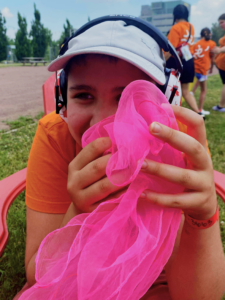 Jeune souriante derrière un foulard rose de jonglerie, assise à l'extérieur.