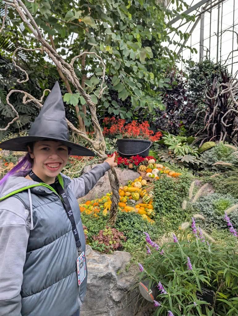 Jeune souriante, déguisée en sorcière pour l'Halloween au Jardin botanique, pointant un décor de chaudron de sorcière.
