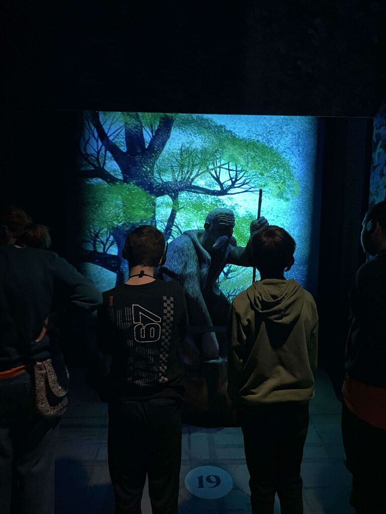 Jeunes avec des audios-guides faisant face à une statue d'un homme âgé, avec une longue barbe, avec un fond de forêt et d'arbres. 