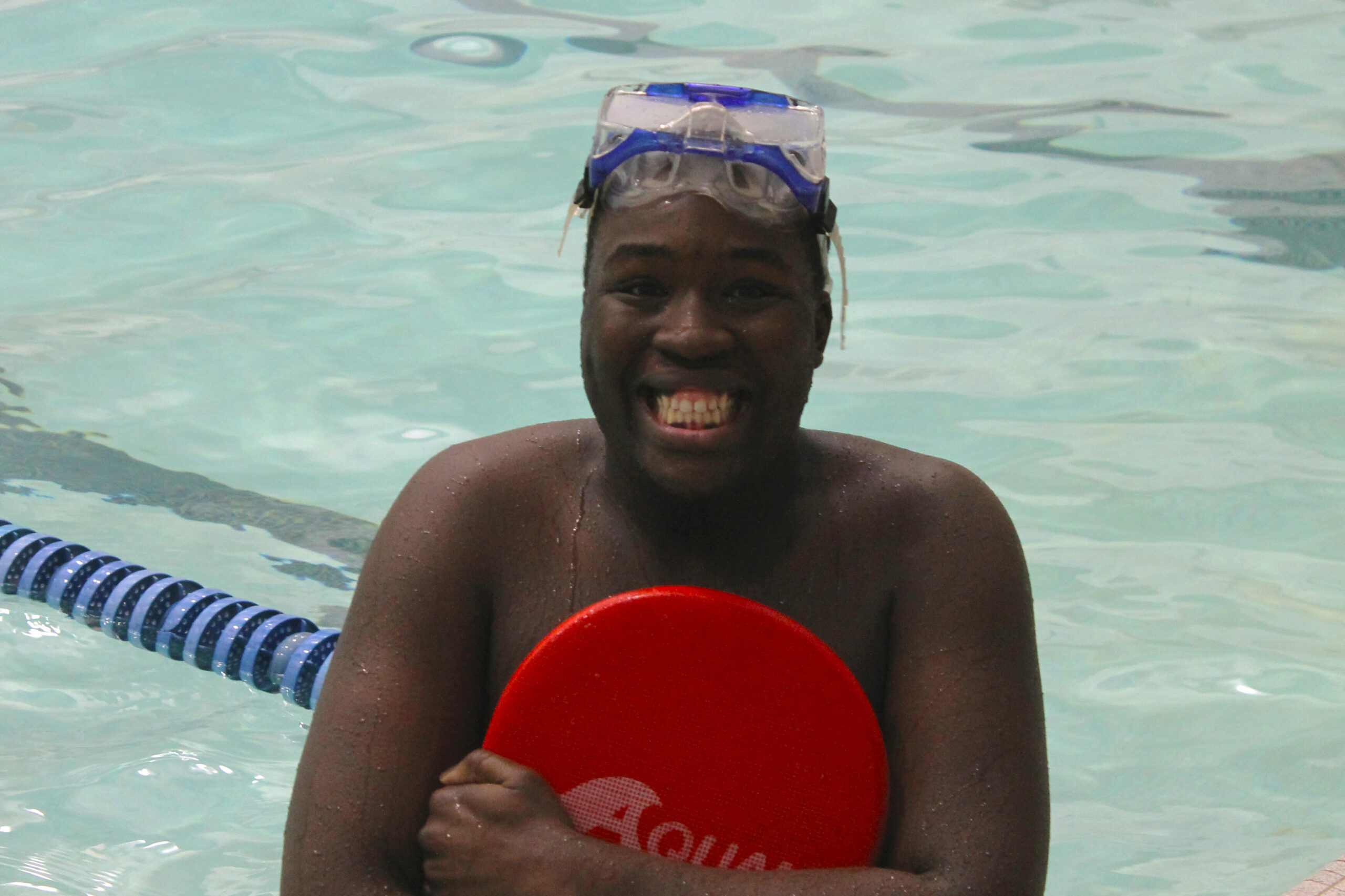 Jeune dans la piscine serrant une planche pour nager, souriant grandement devant la caméra.