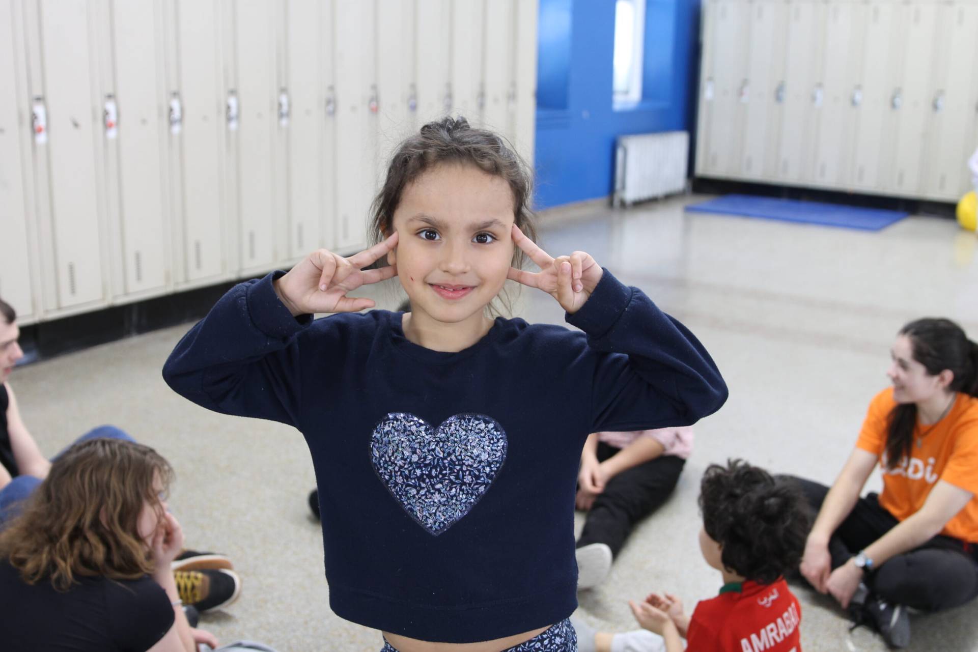  Jeune faisant un signe de paix devant un groupe de personnes assises au sol dans une salle de type scolaire ou récréative.